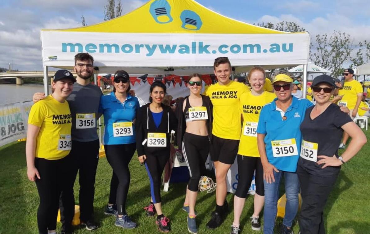 A group of people in sports clothes stand in a field in front of a marquee for the Memory Walk and Jog.