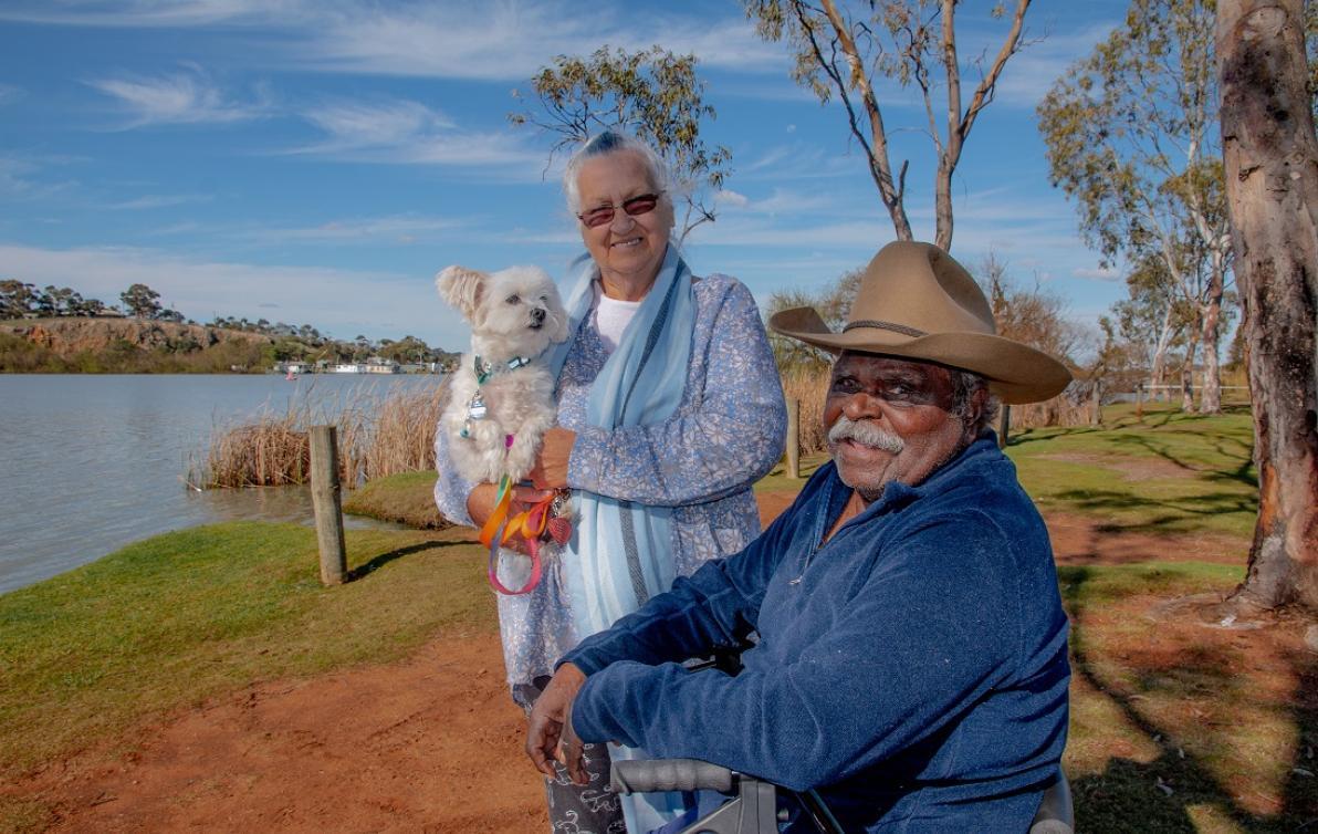 A male and female standing by a lake. Female is holding a small white dog.