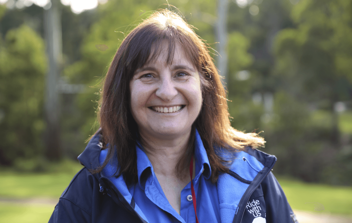 A woman standing outside in the bush smiling at the camera wearing a jacket with a logo that says ride with us