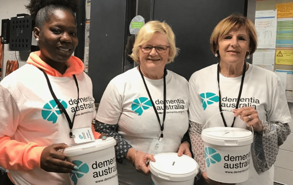 Three people wearing Dementia Australia t-shirts are holding cash collection buckets at a fundraising event.