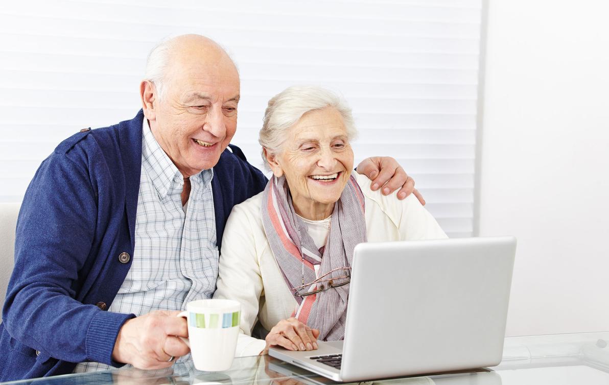 A couple smiling and looking at a laptop on their table. Man has his arm around her.