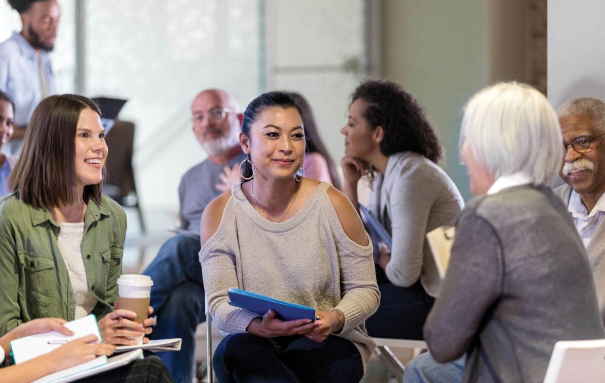 A group of people sit in a circle of chairs, holding notepads and coffees, talking animatedly.