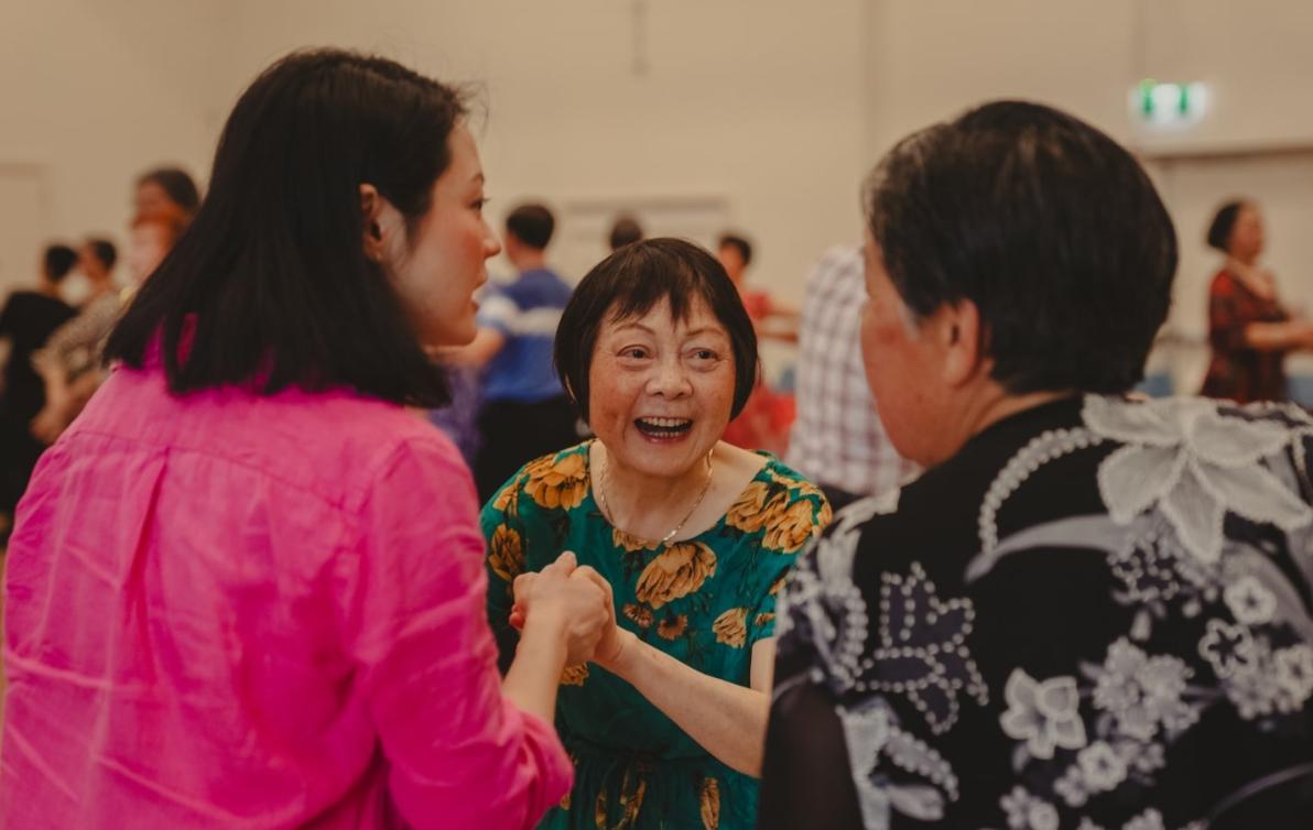 A woman stands smiling in a hall. Two other people hold her hands and talk to her as others gather behind.