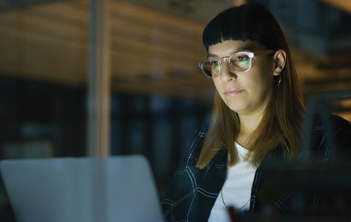 A woman sits before a laptop. The screen is slightly reflected in her glasses.