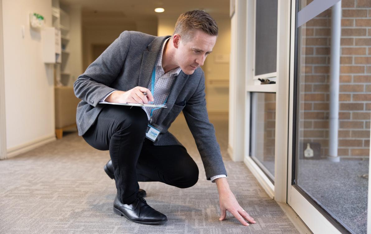 A man with a clipboard inspects the carpet of a room.