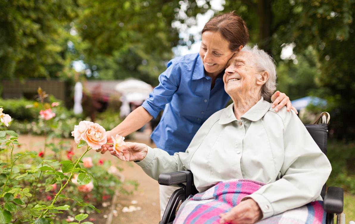 An elderly female in a wheelchair being hugged by a younger female in a garden, both smiling