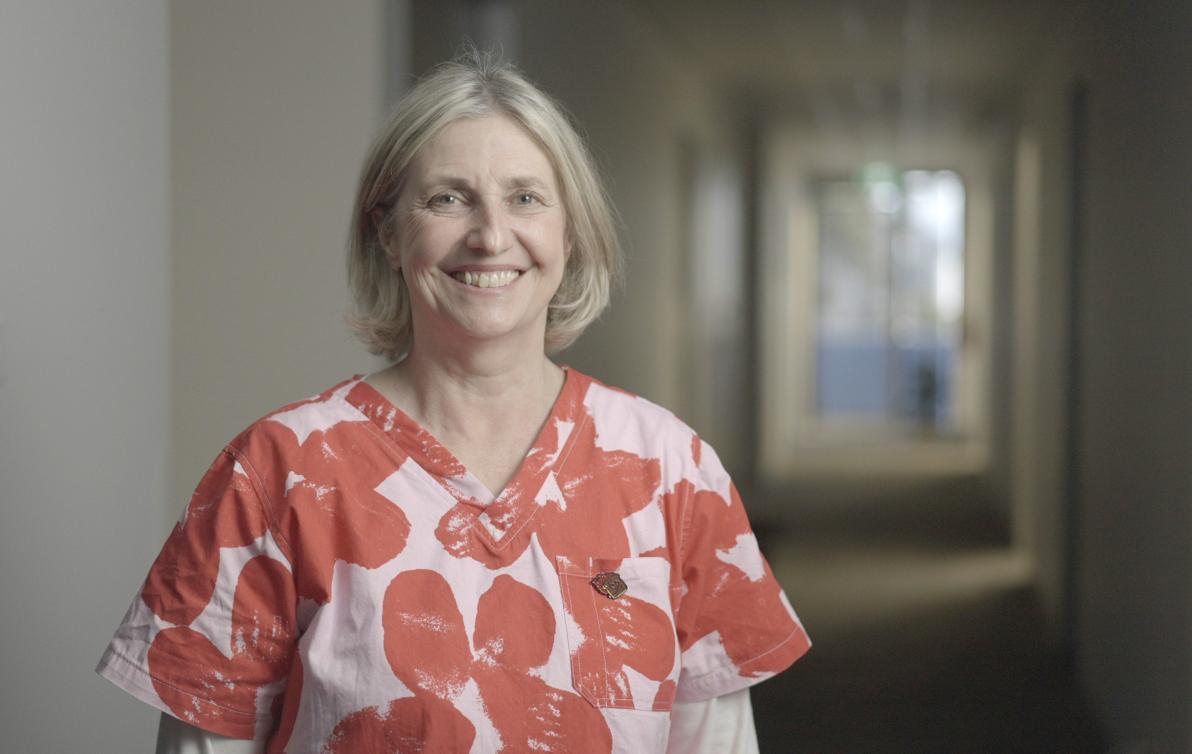 A woman wearing colourful scrubs stands in the hallway of a hospital smiling