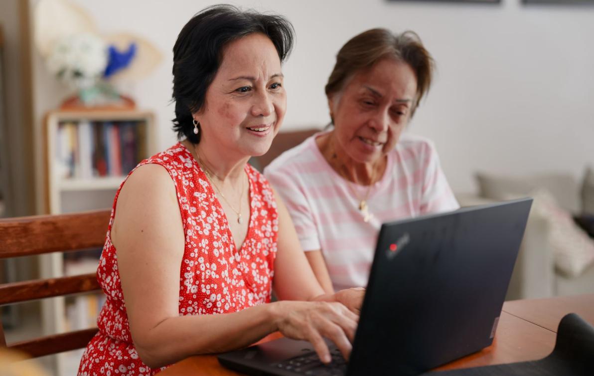 Two women sit at a table, smiling and looking at a laptop screen.