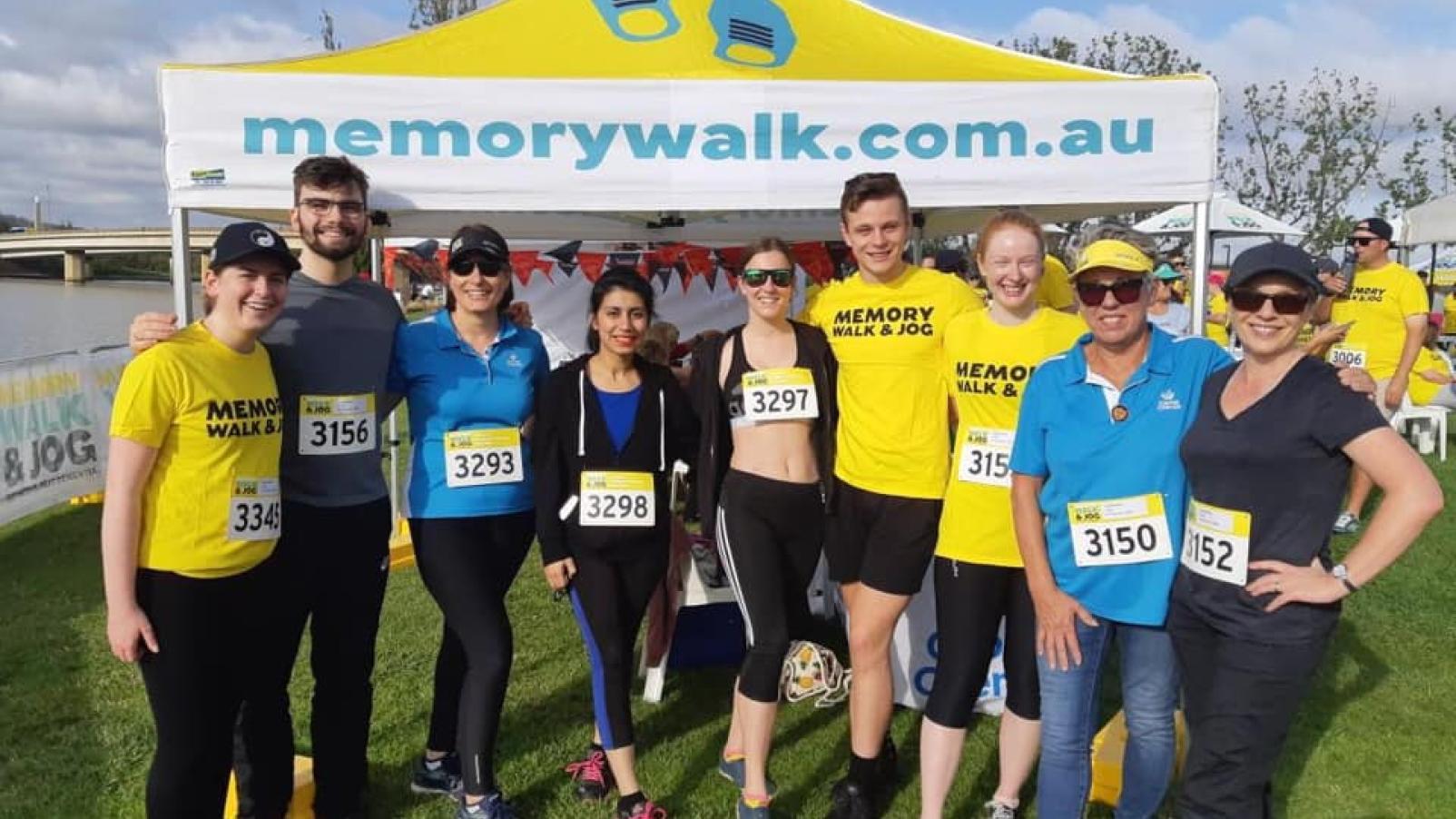 A group of people in sports clothes stand in a field in front of a marquee for the Memory Walk and Jog.