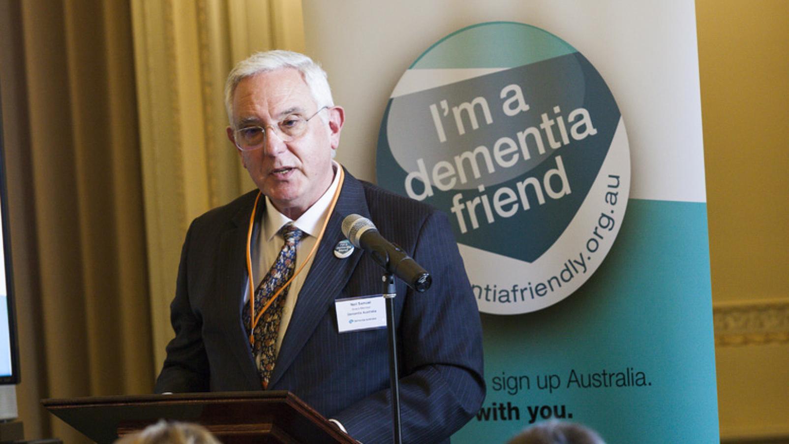 Neil Samuel, a man wearing a suit and glasses, stands at a lectern and speaks to an audience.