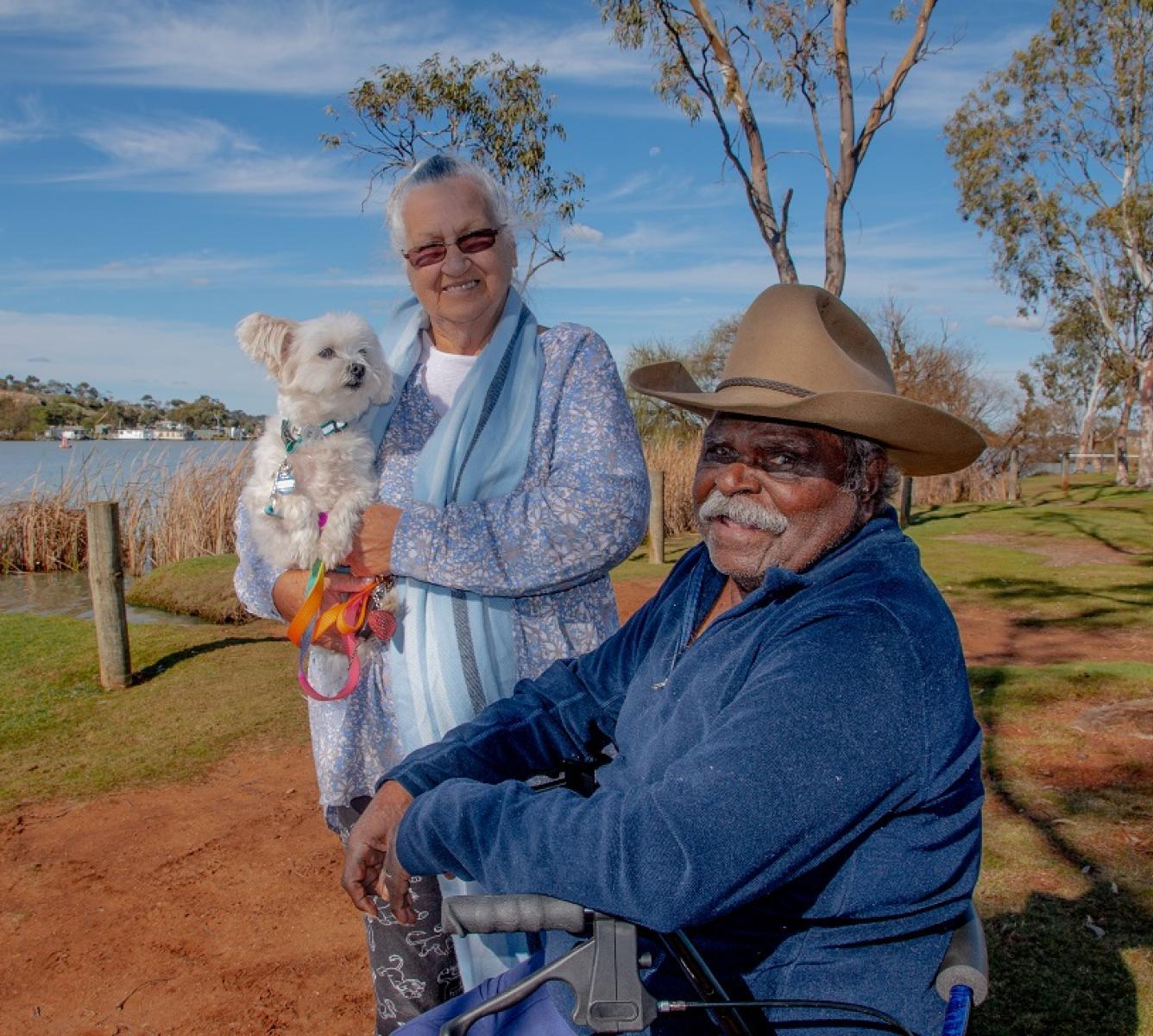 A male and female standing by a lake. Female is holding a small white dog.