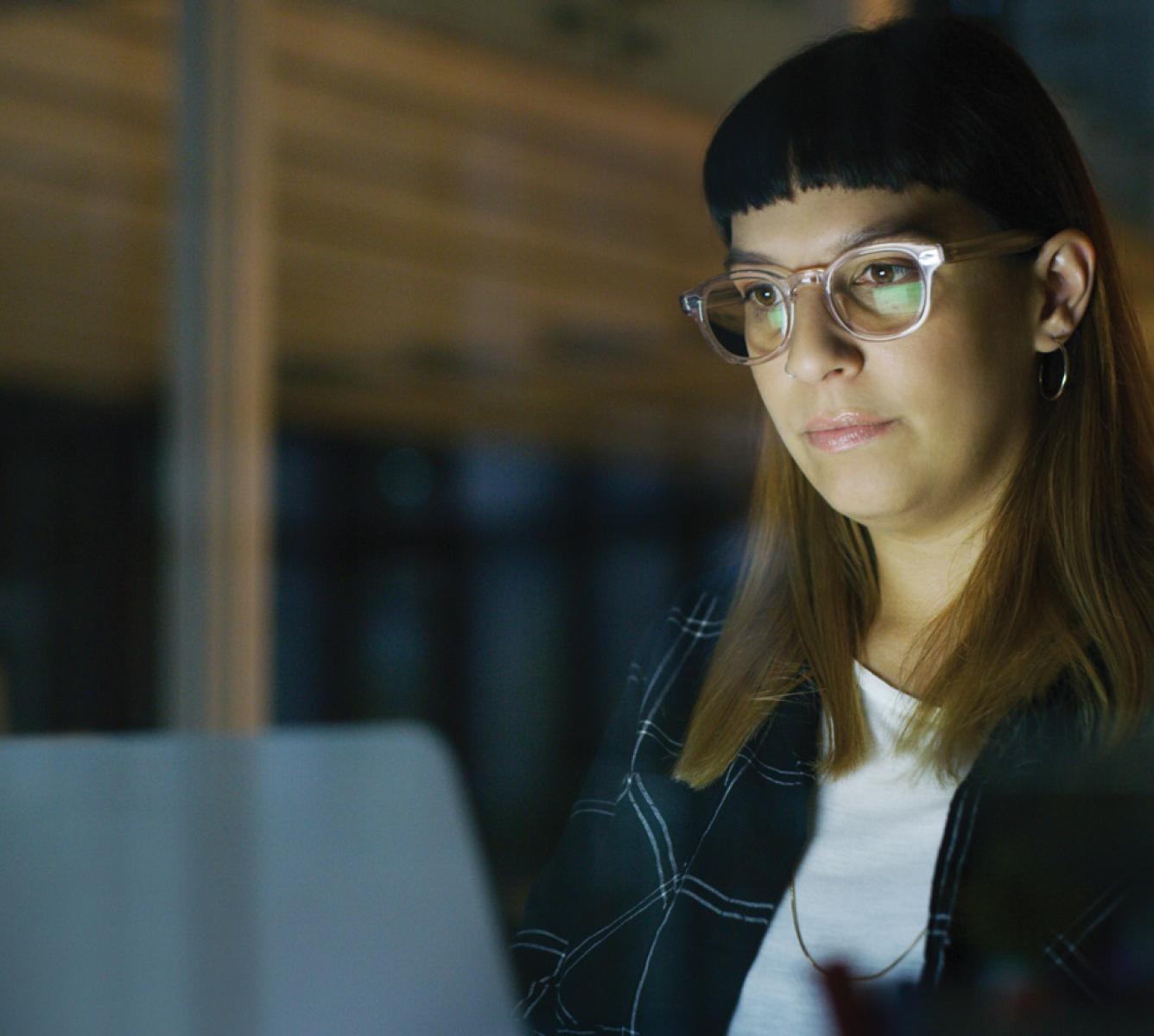 A woman sits before a laptop. The screen is slightly reflected in her glasses.