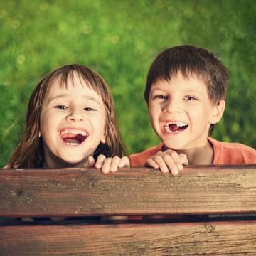 A yong girl and young boy smiling at the camera from behind a park bench